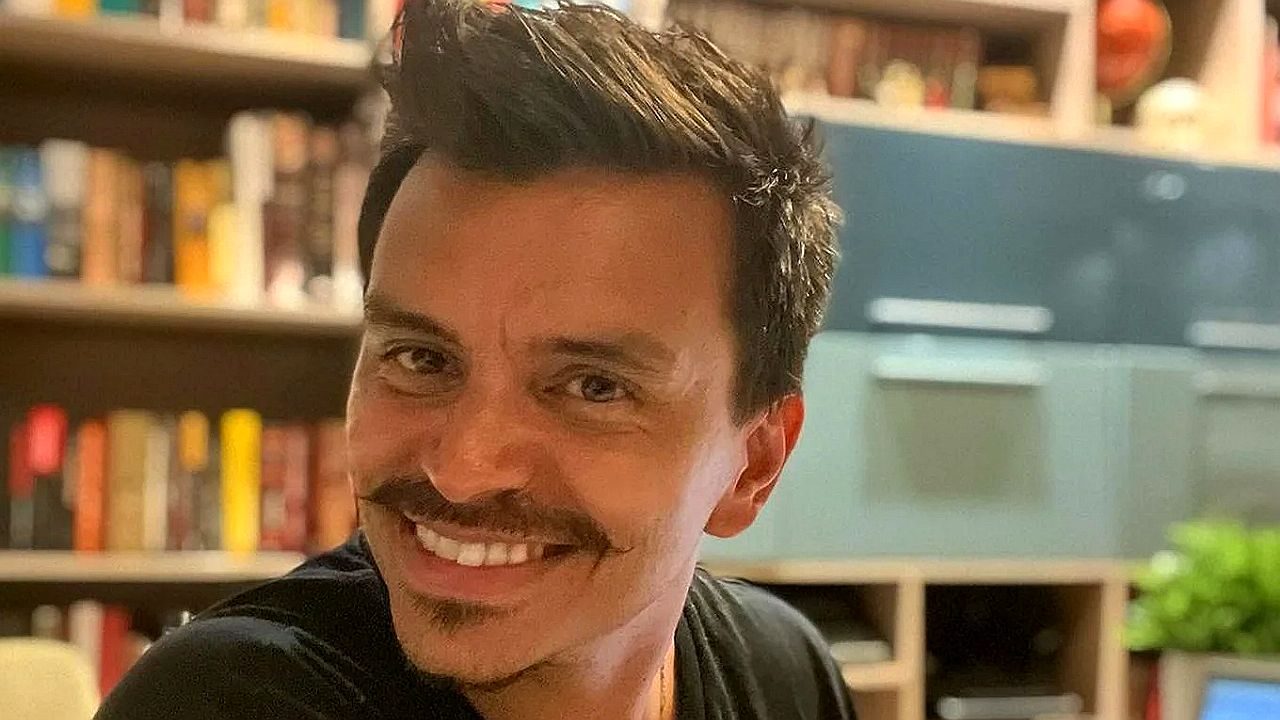 Homem com bigode sorri para a câmera em ambiente interno com estante de livros ao fundo