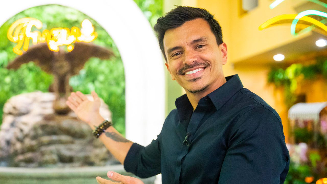 Autor Gustavo Reiz usando camisa preta e pulseiras, gesticulando com a mão direita em frente a uma fonte decorativa