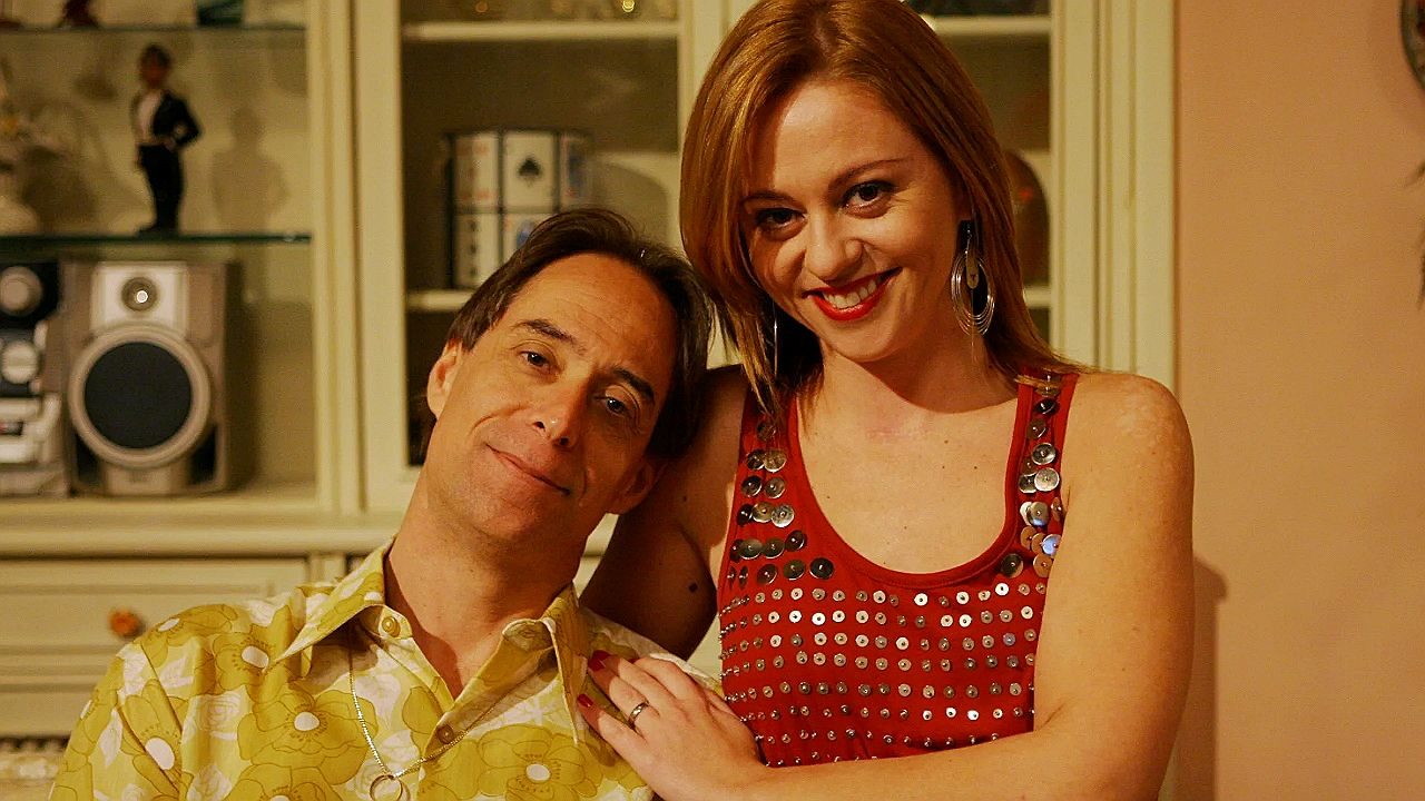 Casal sorri junto em sala de estar com estante ao fundo em cena de série ou filme