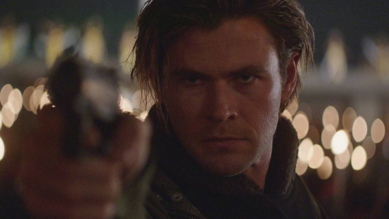 Homem com expressão intensa aponta uma arma diretamente para a câmera em um ambiente noturno iluminado por luzes desfocadas ao fundo, transmitindo tensão e clima de confronto.