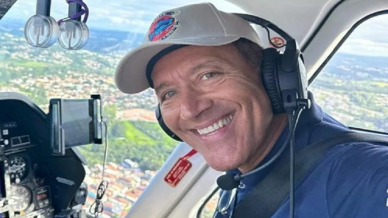 Comandante Hamilton sorrindo dentro de uma aeronave, usando boné claro, fones de ouvido de piloto e uniforme azul, com vista aérea de uma cidade ao fundo