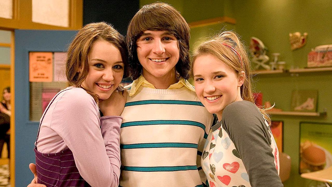 Mitchel Musso entre Miley Cyrus e Emily Osment sorrindo em cena da série Hannah Montana