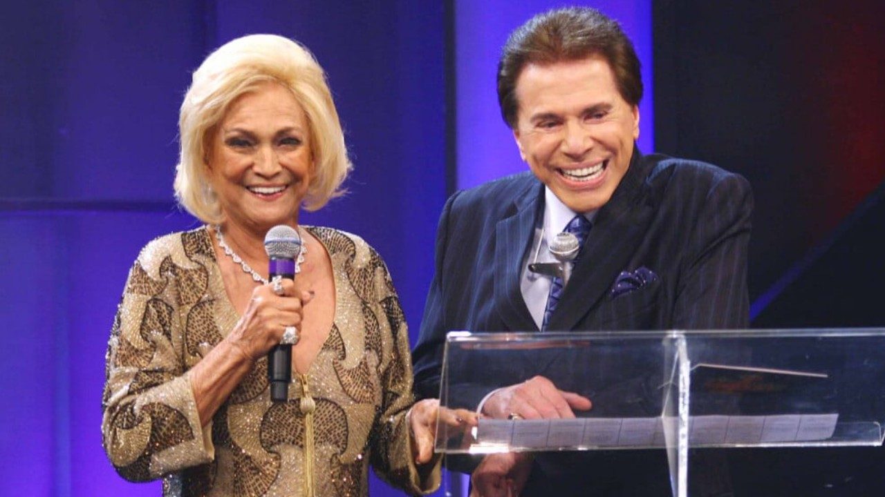 Hebe Camargo de vestido dourado segura microfone ao lado de Silvio Santos em palco com fundo azul
