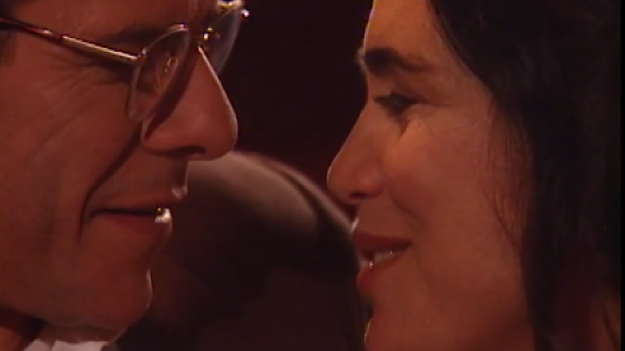 A imagem captura um momento de grande intimidade e cumplicidade da personagem Helena, interpretada por Regina Duarte, na novela História de Amor (1995). Ela está próxima de um homem, ambos sorrindo suavemente, em um clima de romance e conexão emocional.