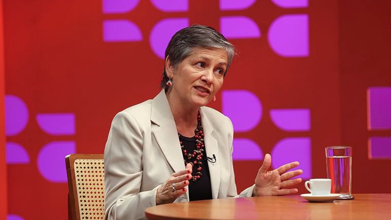 Mulher de cabelo curto fala gesticulando durante entrevista em estúdio com mesa e copo de água