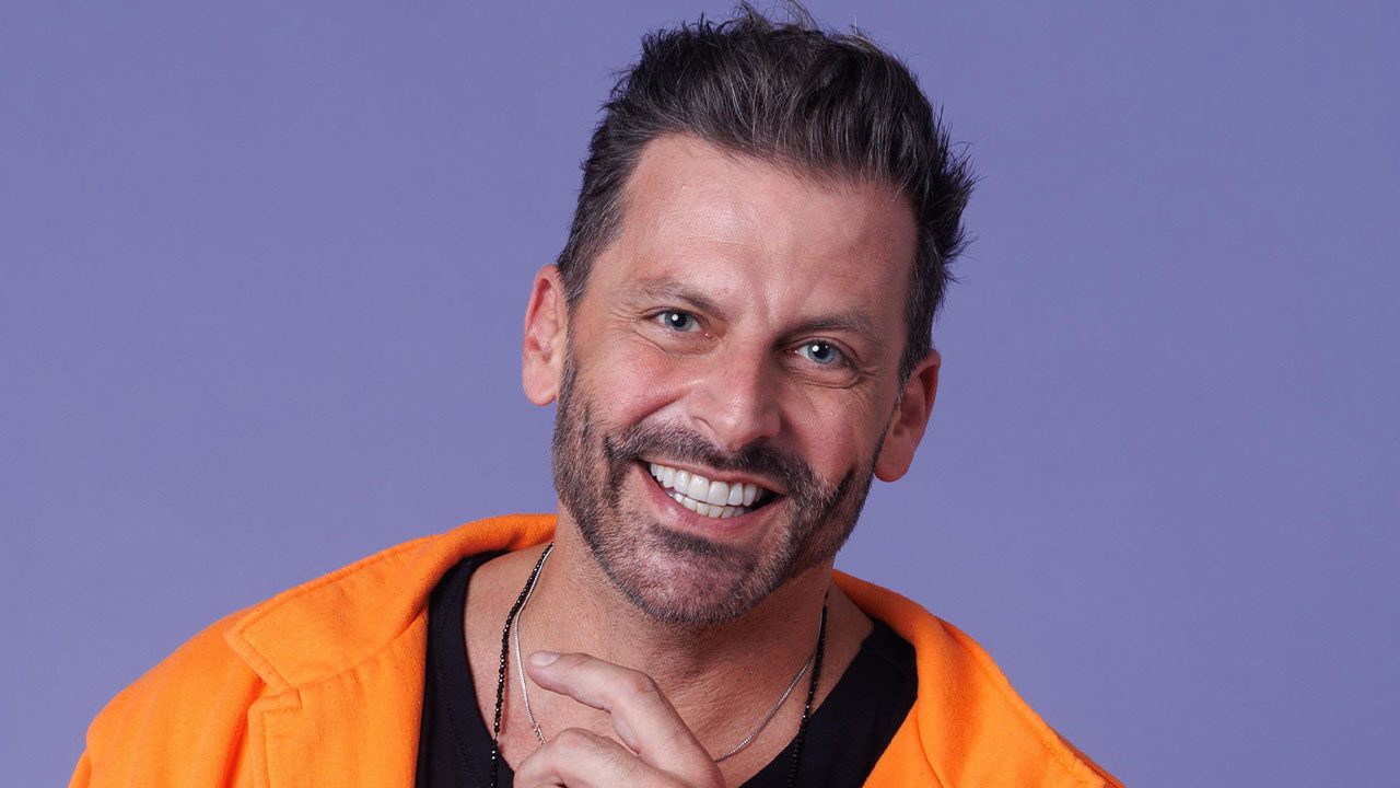 Henri Castelli sorrindo com blazer laranja sobre camiseta preta em frente a fundo lilás