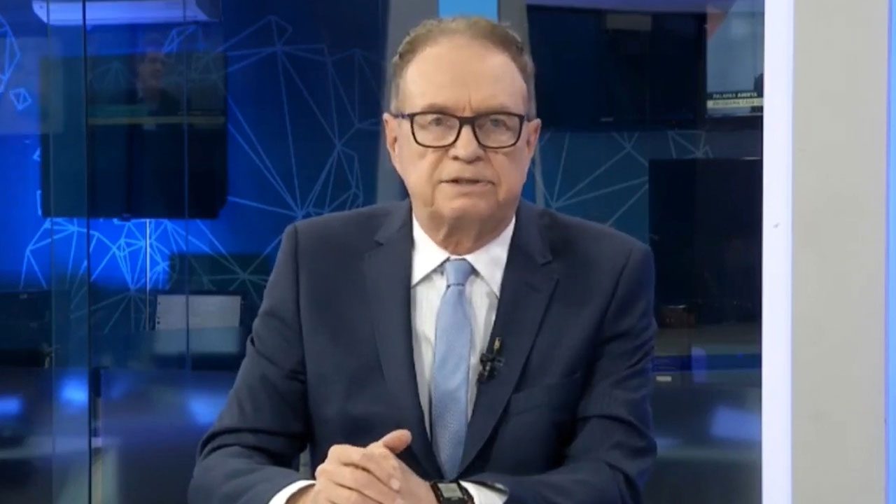 Hermano Henning de terno azul e gravata clara apresentando telejornal no estúdio da Rede Brasil de Televisão