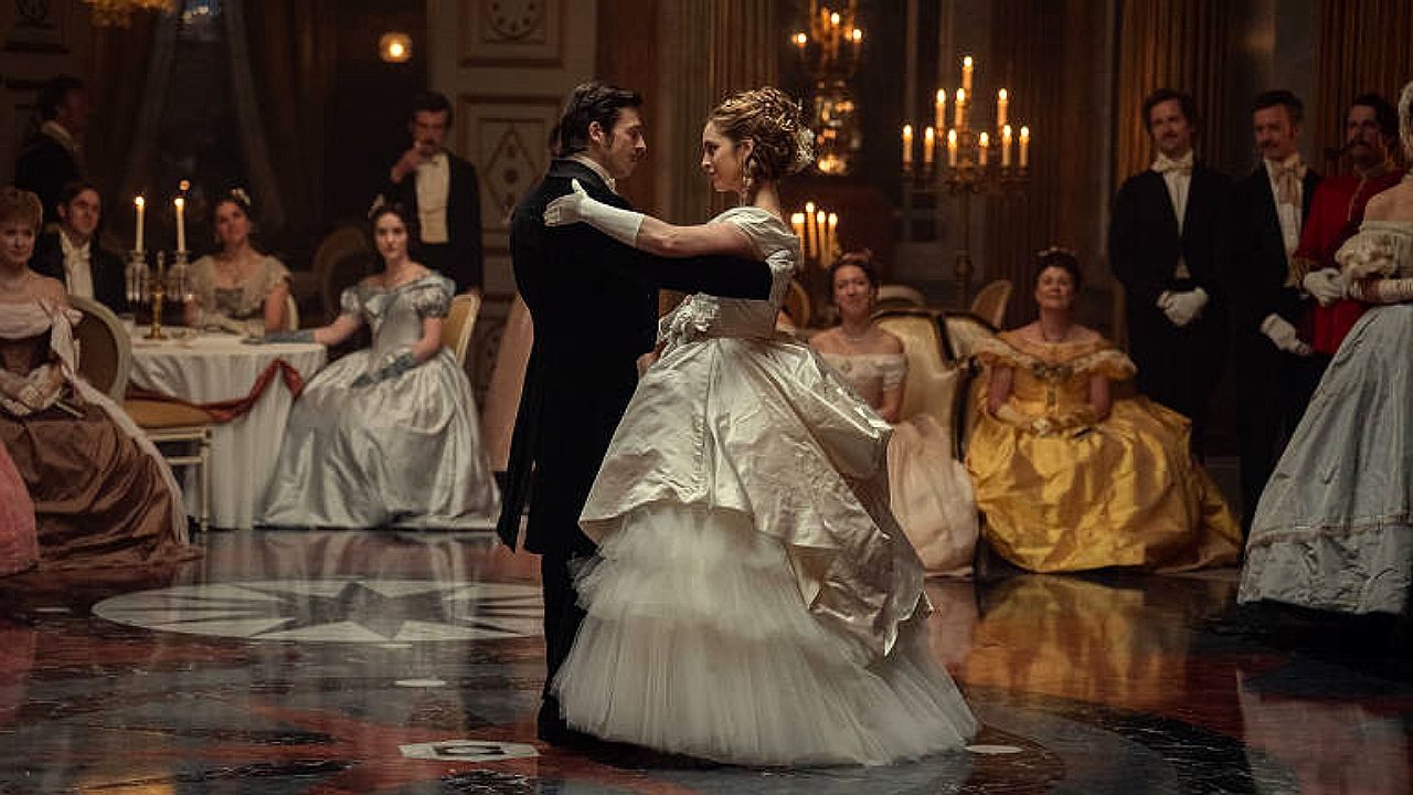 Casal dança em salão iluminado por velas durante baile da alta sociedade em cena de drama de época