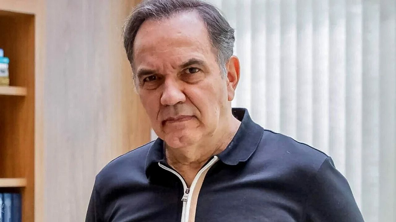 Homem de camisa polo preta olha sério para a câmera em ambiente interno com estante ao fundo