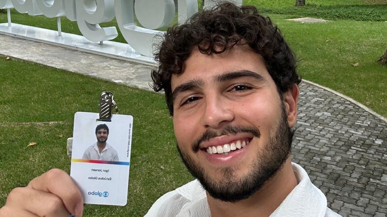 Igor Jansen sorri segurando crachá da Globo em selfie feita na área externa da emissora