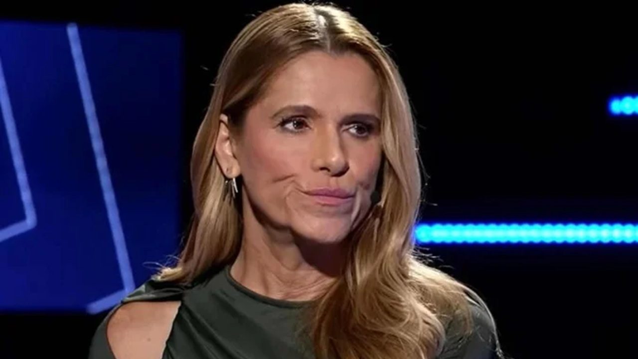 Mulher loira de expressão séria, usando vestido verde de ombro vazado e microfone preso ao rosto, aparece em estúdio escuro com luzes azuis ao fundo, durante o que parece ser uma gravação ou entrevista.