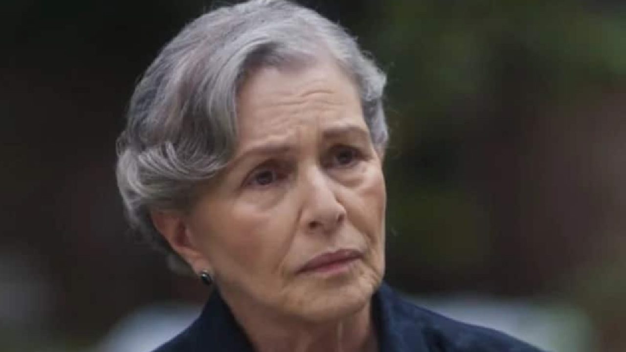 Mulher idosa com cabelos grisalhos curtos e expressão séria aparece em cena externa, sob luz natural e com fundo desfocado