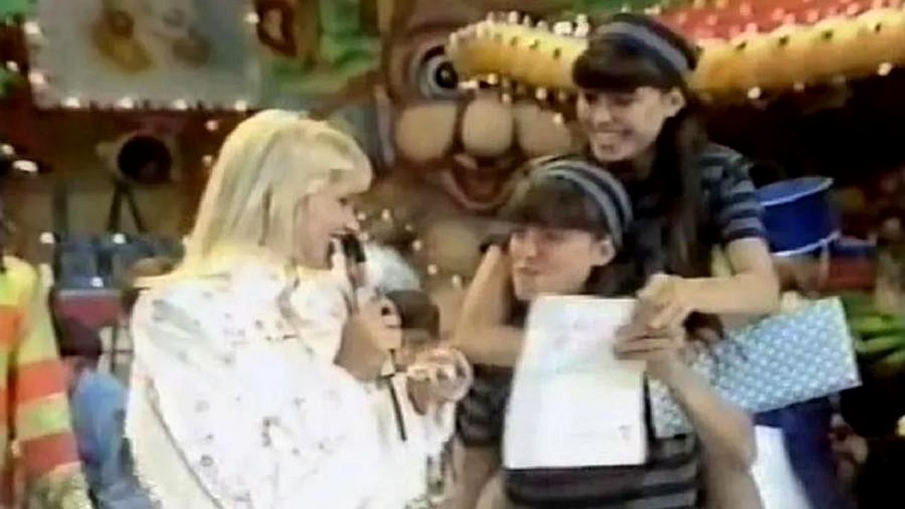 Apresentadora loira entrevista duas meninas em palco infantil com cenário colorido ao fundo durante programa de TV