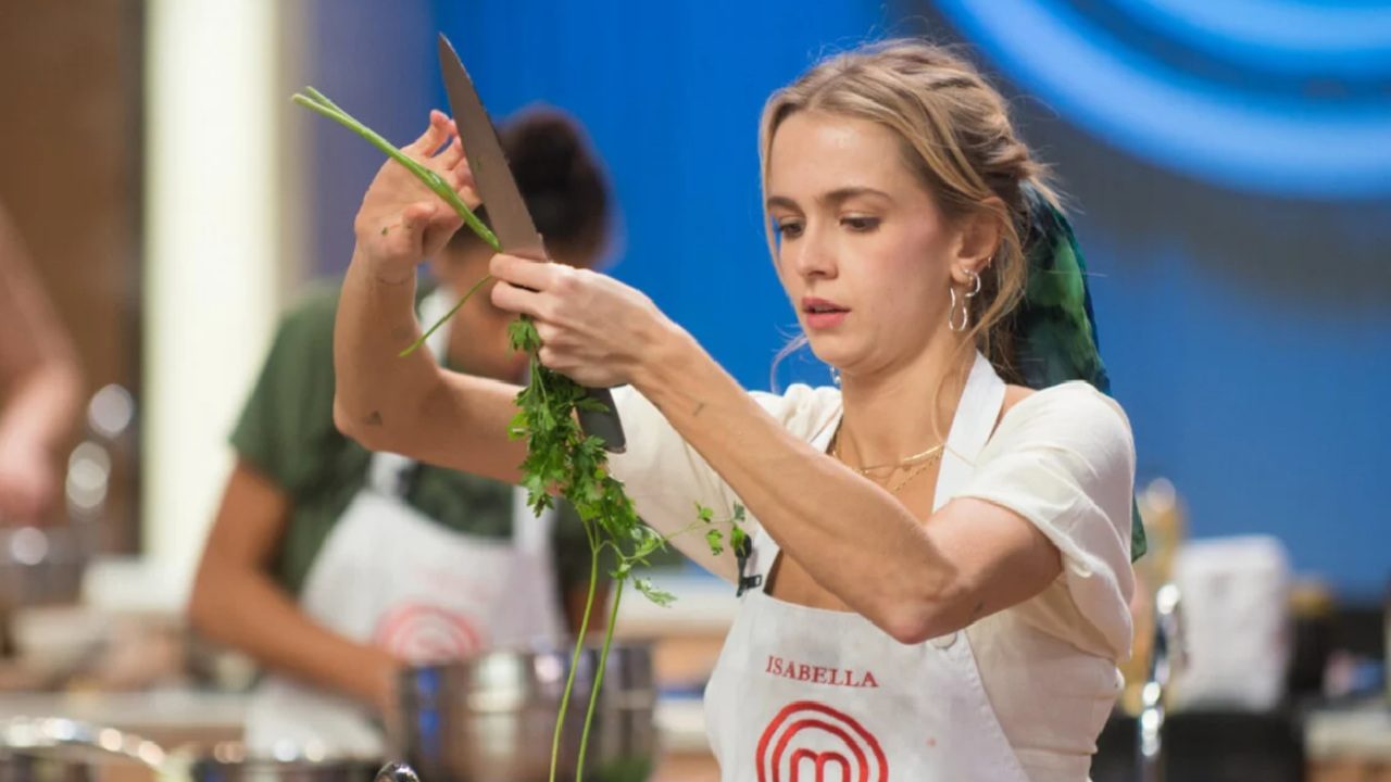 Mulher com avental do MasterChef segura uma faca e corta ervas frescas durante prova culinária no estúdio do programa