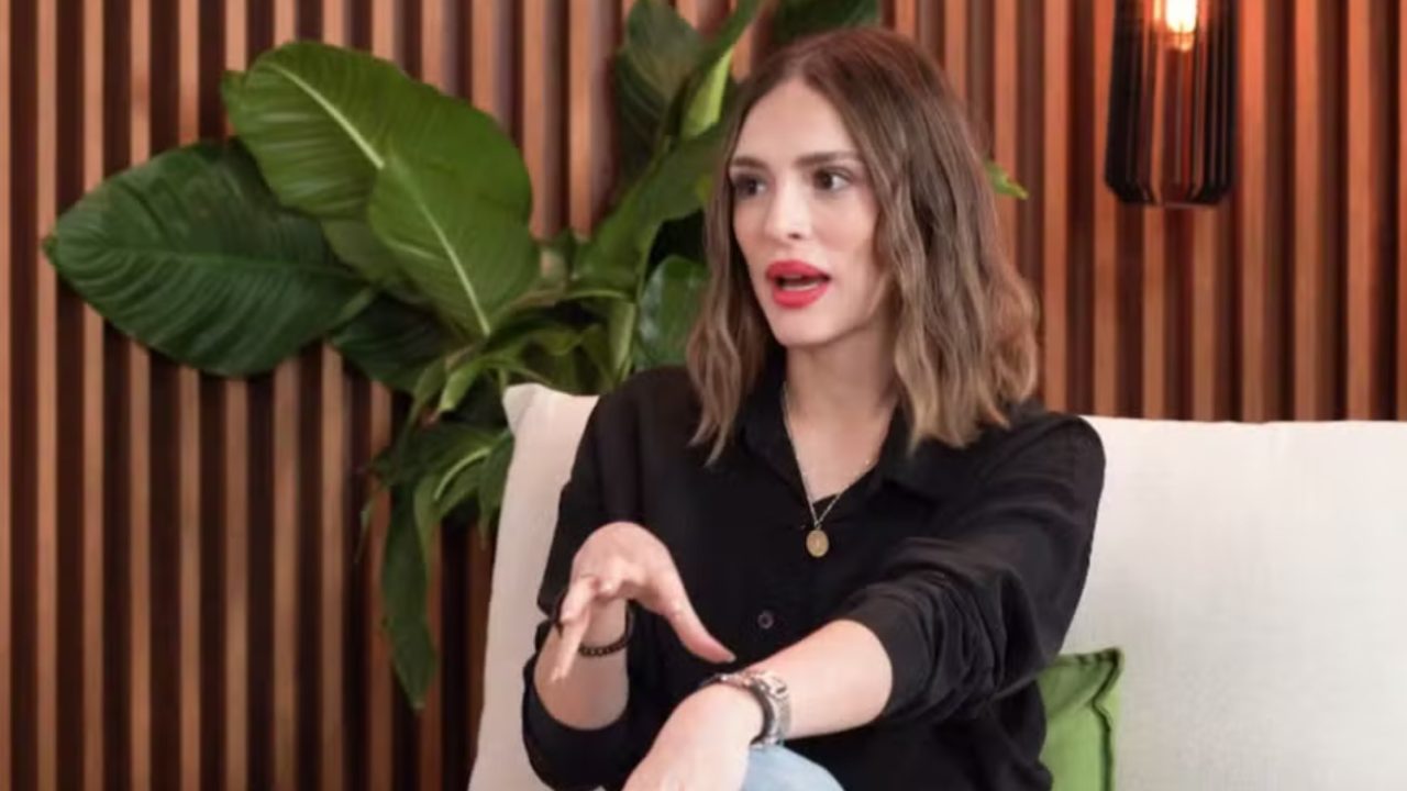 Mulher sentada em sofá claro gesticula enquanto fala, em ambiente interno com parede de ripas de madeira e plantas ao fundo, sugerindo clima de entrevista ou conversa informal.