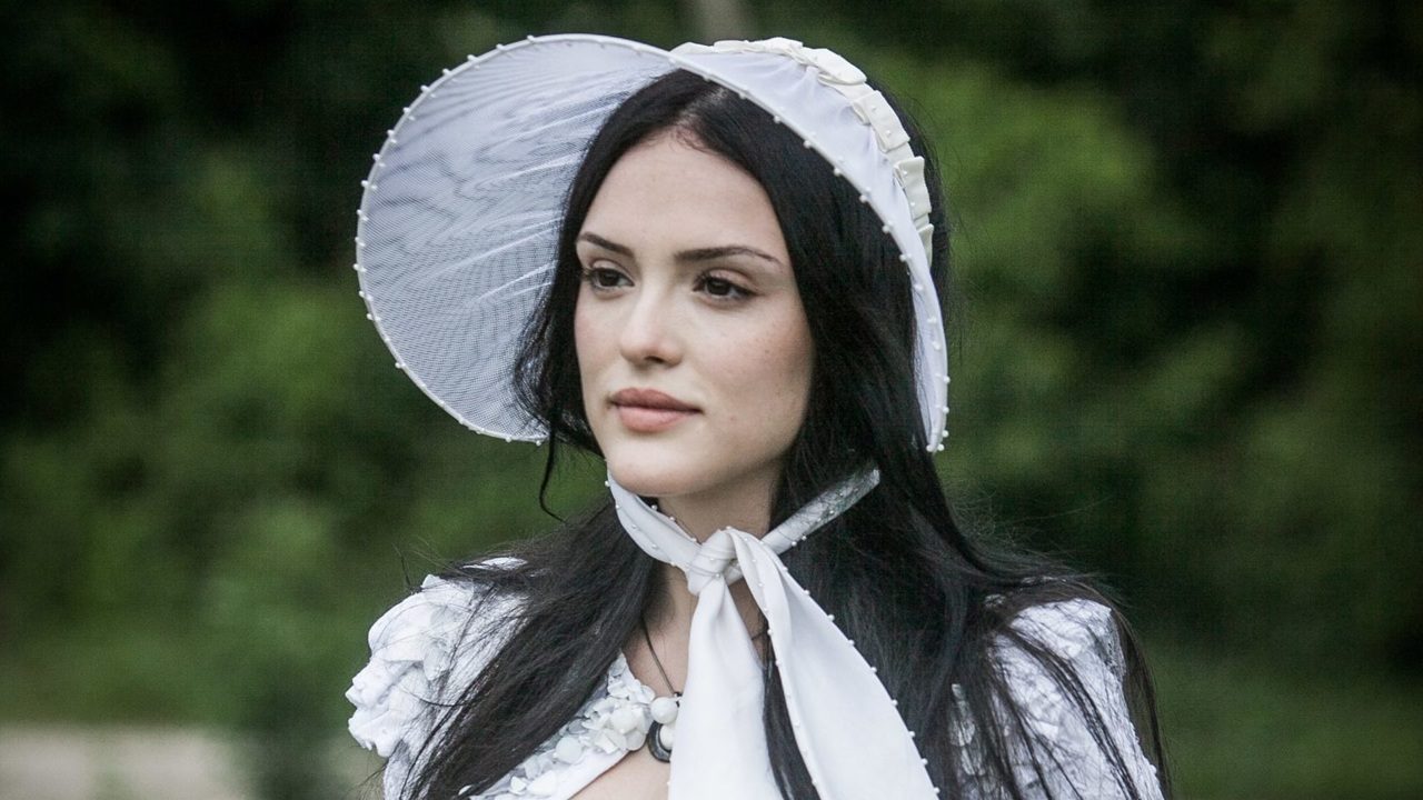 Mulher jovem com longos cabelos pretos aparece ao ar livre, vestindo um chapéu branco estilo camponês com detalhes delicados e laço sob o queixo. Ela usa roupa branca com adornos florais e olha serenamente para a frente; ao fundo há vegetação esverdeada desfocada