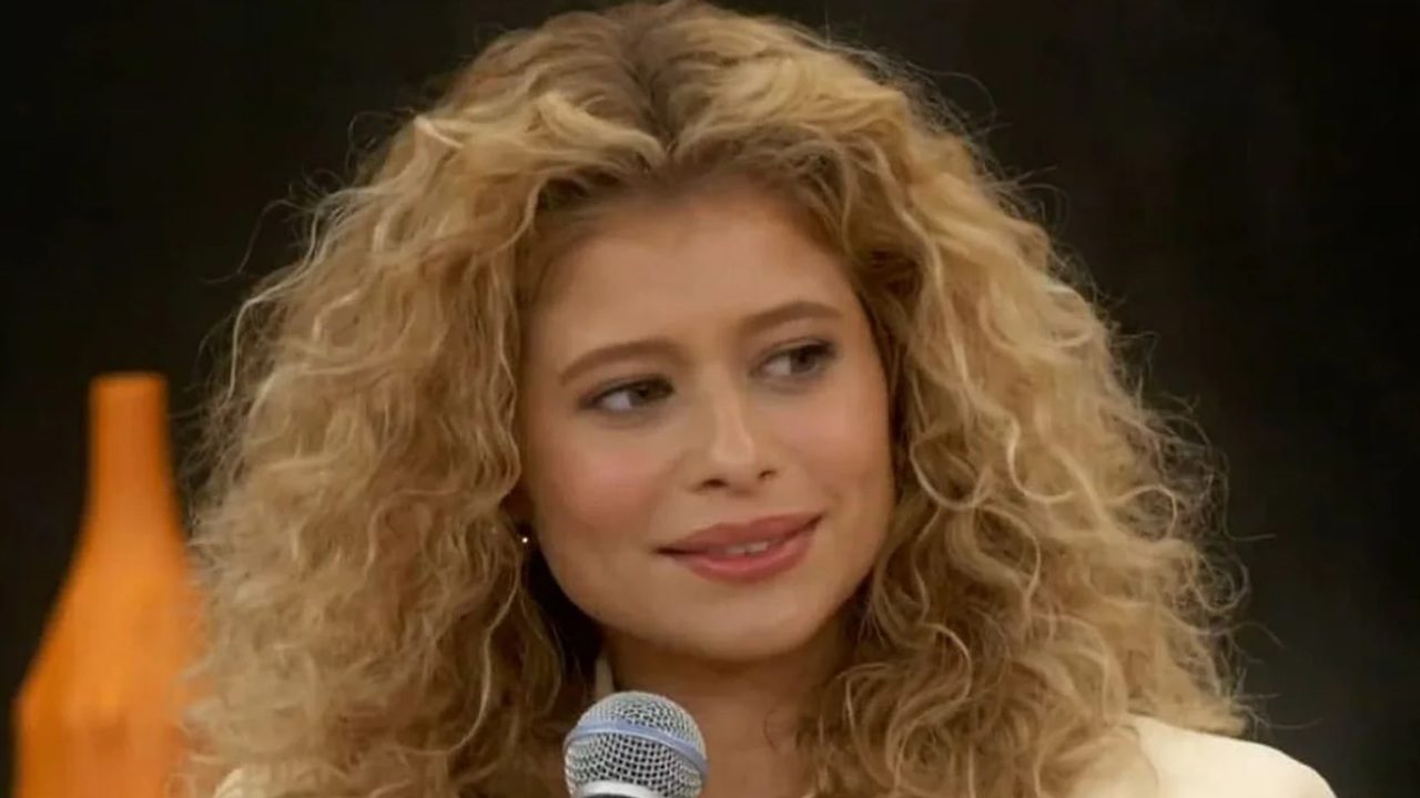 Mulher de cabelos loiros cacheados segura microfone e sorri durante entrevista em estúdio