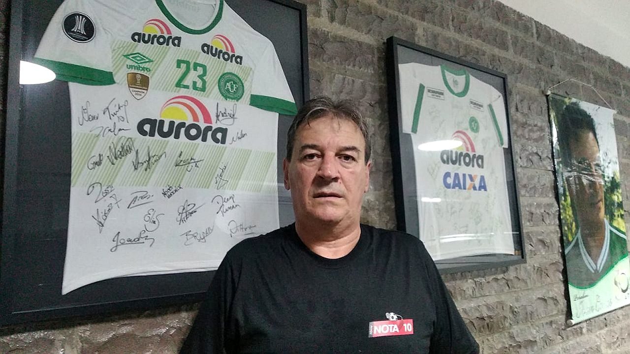 Ivan Carlos posa diante de camisas autografadas da Chapecoense em ambiente com decoração dedicada ao clube