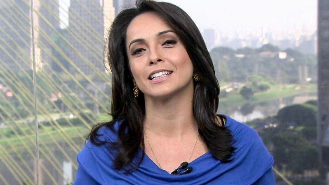 Izabella Camargo de blusa azul sorri diante de janela com vista urbana durante apresentação em estúdio de TV