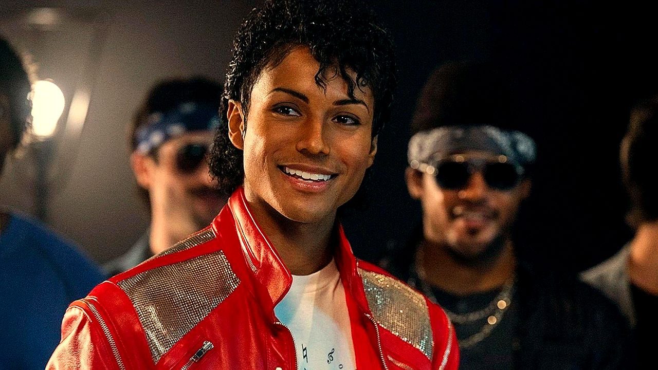 Texto Alternativo disse: Cantor caracterizado como Michael Jackson com jaqueta vermelha em cena de performance musical com dançarinos ao fundo