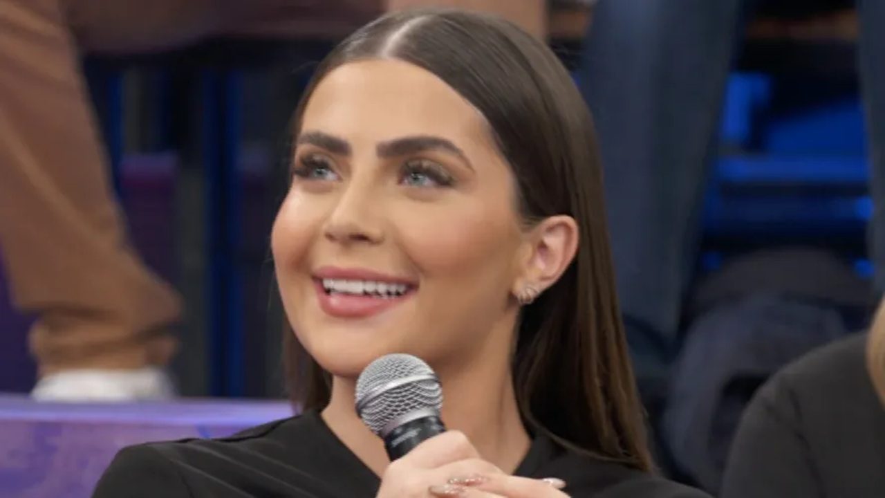 Mulher sorrindo enquanto segura um microfone durante participação em programa de TV, com maquiagem destacada e cabelo liso dividido ao meio; ao fundo, público desfocado em ambiente de estúdio.