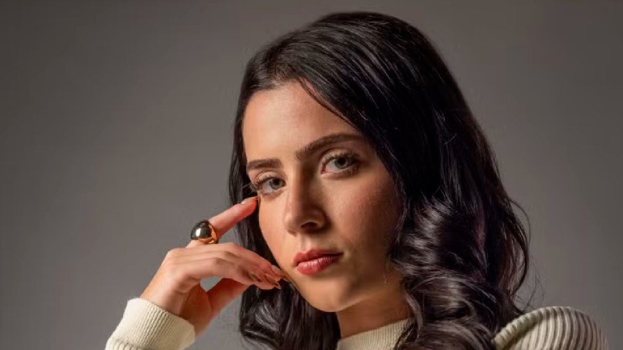 Mulher de cabelos escuros e cacheados posa com expressão séria, apoiando o rosto na mão. Ela veste uma blusa clara e usa um anel grande dourado, em frente a um fundo neutro e iluminado suavemente, transmitindo um ar introspectivo e elegante