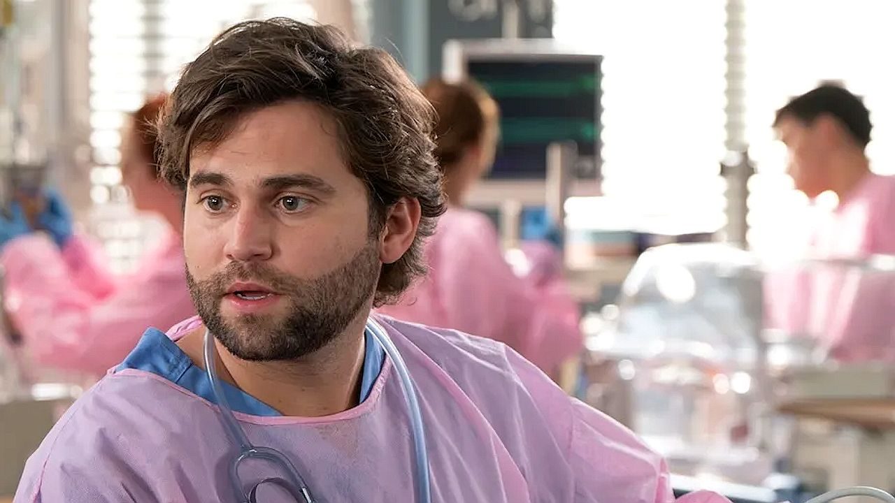 Médico em Grey's Anatomy com estetoscópio usa uniforme hospitalar rosa e conversa em enfermaria com equipe ao fundo