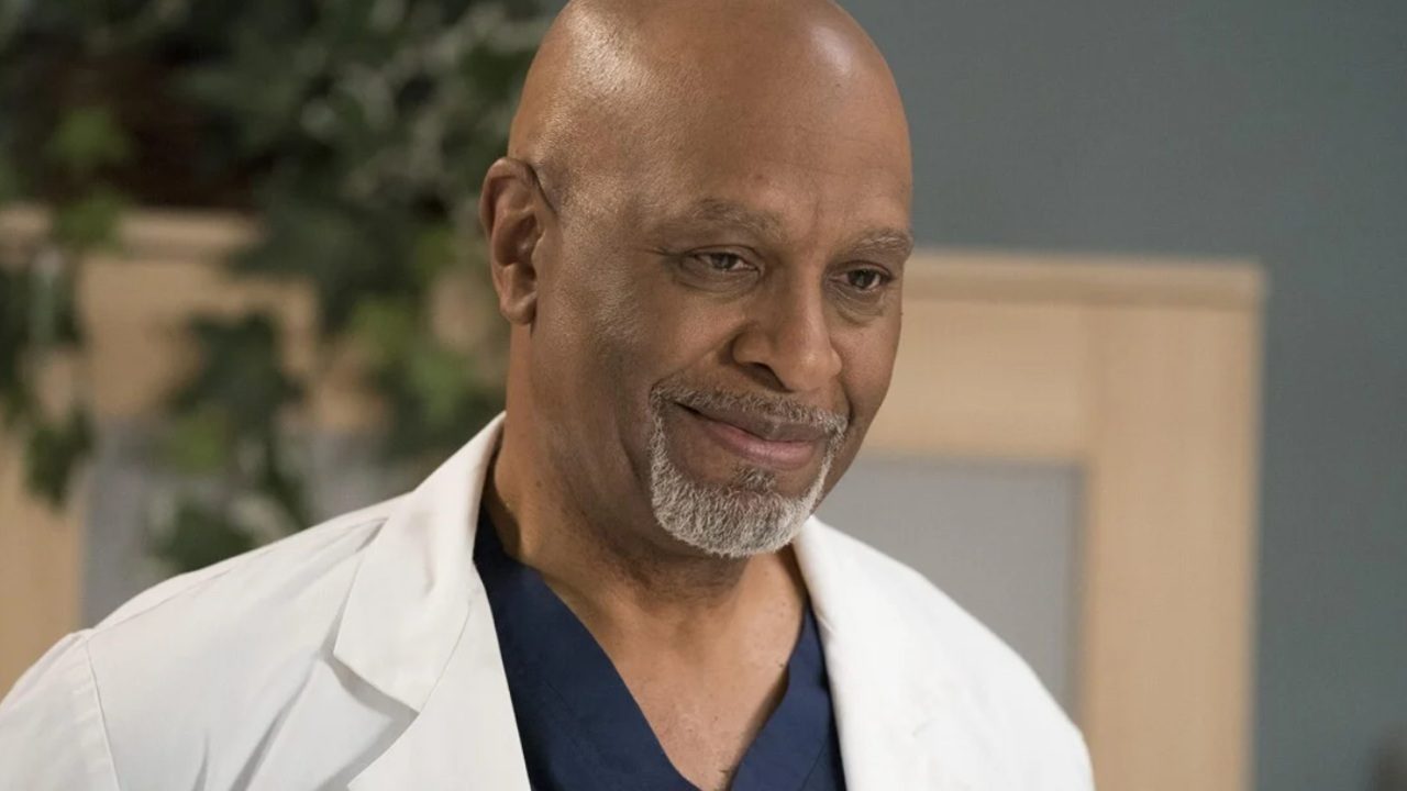 A imagem mostra um homem calvo com barba grisalha, usando jaleco branco e roupa médica azul por baixo, sugerindo que ele é um médico