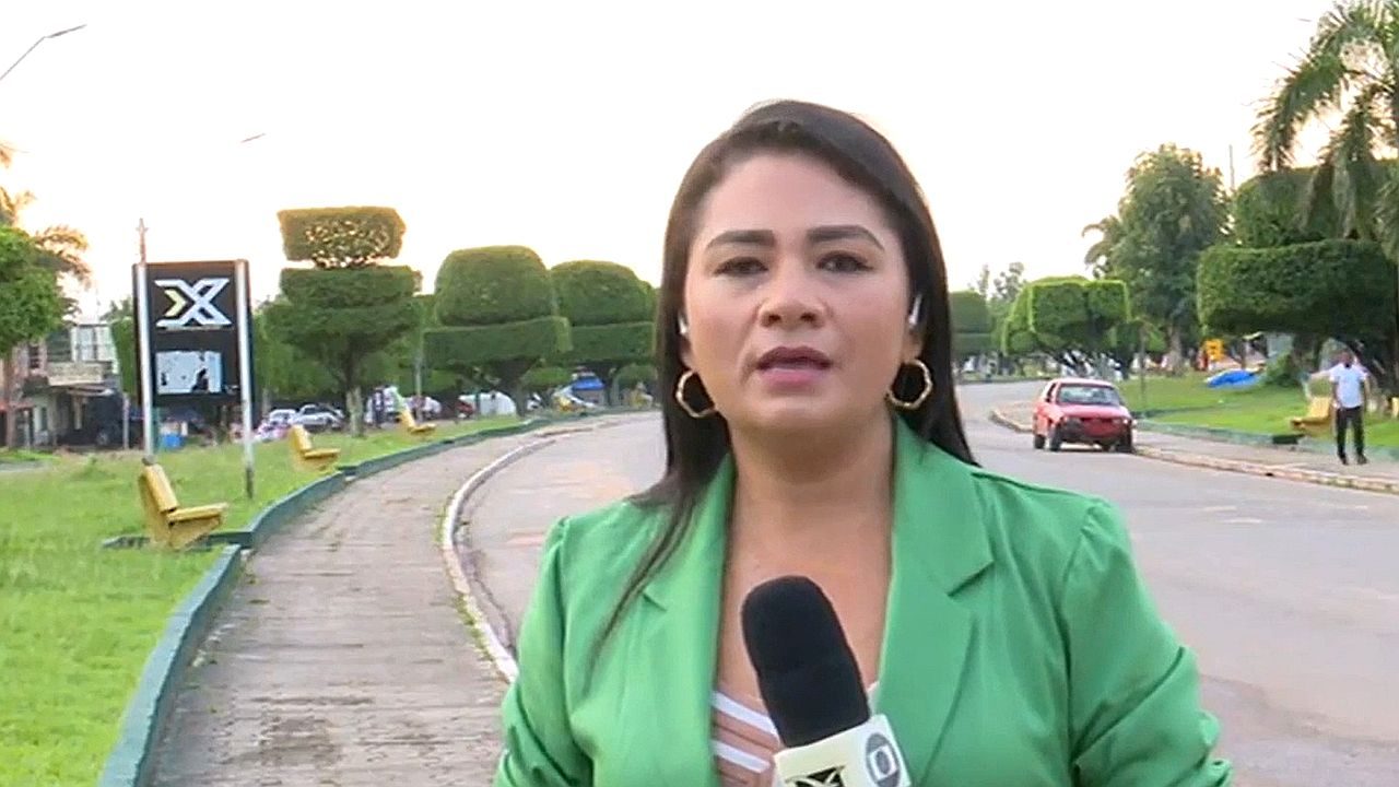 Uma mulher aparece em pé à beira de uma rua, segurando um microfone, como em uma reportagem externa. Ela usa blazer verde e brincos, com expressão séria e focada, olhando diretamente para a câmera. Ao fundo, há uma via com carros passando, bancos ao longo da calçada e árvores podadas em formato geométrico, sugerindo um ambiente urbano organizado.