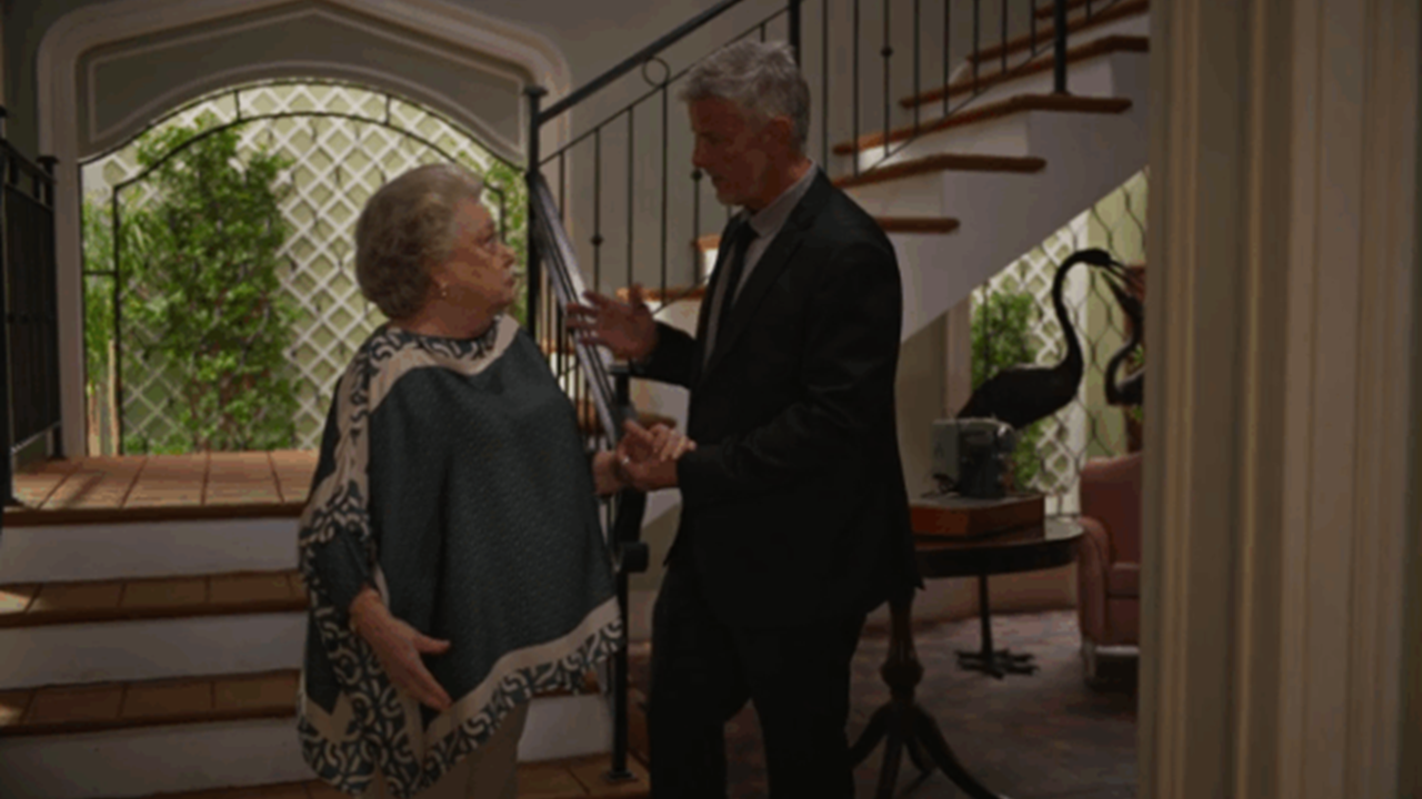 Uma mulher idosa e um homem de cabelos grisalhos conversam em um hall de entrada iluminado, próximo a uma escada com grades pretas. A mulher usa uma capa estampada em tons de verde e bege, enquanto o homem veste terno escuro e segura a mão dela. Ao fundo, há plantas, uma parede com treliça branca e parte de uma sala com móveis