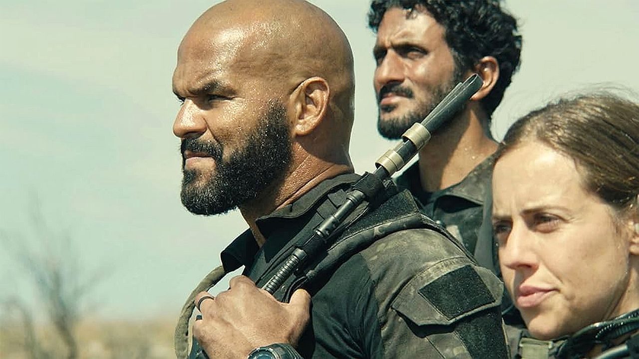 Três pessoas em trajes táticos estão ao ar livre, sob luz forte do sol, com expressão séria e atenta. Em primeiro plano, um homem careca com barba segura um rifle apoiado no ombro, vestindo uniforme escuro e relógio no pulso. Atrás dele, outro homem com cabelo cacheado e barba observa ao longe. À direita, uma mulher também com roupa tática olha na mesma direção. O cenário parece árido, sugerindo missão ou operação em ambiente desértico ou rural.