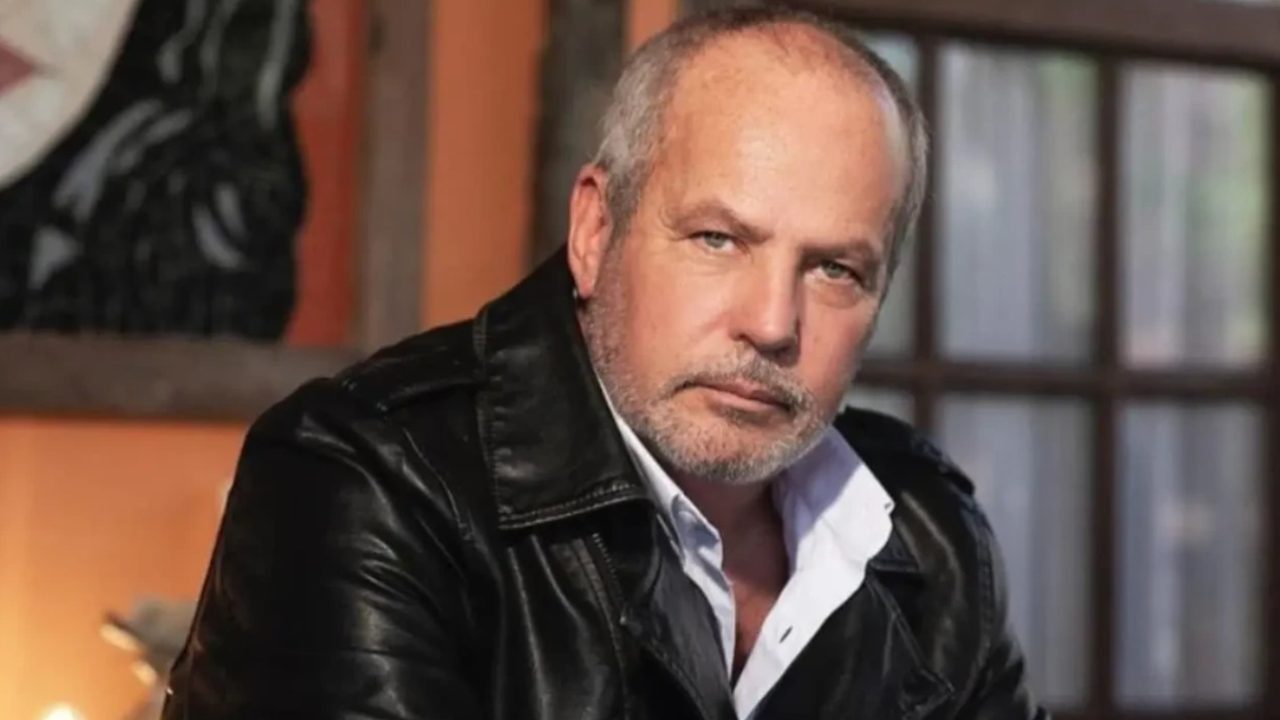 A imagem mostra um homem de expressão séria, vestindo uma jaqueta de couro preta sobre uma camisa branca. Ele está em um ambiente interno com iluminação suave, e ao fundo há uma parede alaranjada com uma pintura e uma janela de vidro. O clima da foto é introspectivo e um pouco dramático.