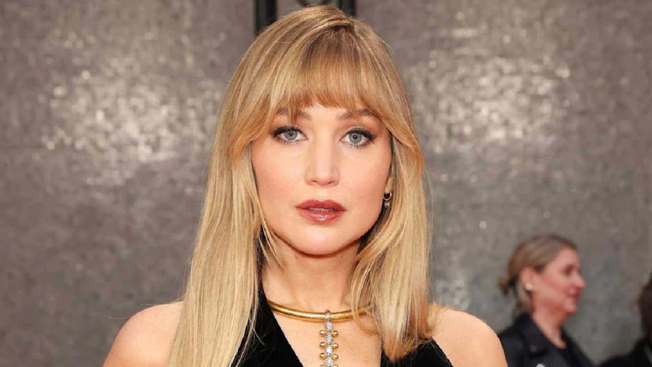 Mulher de cabelos loiros e franja usa vestido preto e colar dourado com pingente em evento com fundo prateado brilhante e pessoas ao fundo desfocadas