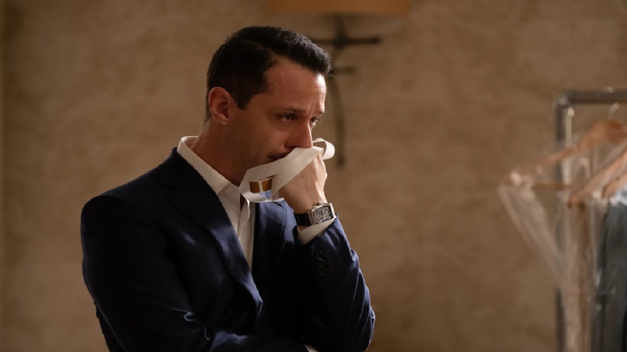 Jeremy Strong em cena com expressão séria, vestindo terno escuro e camisa branca, segurando um lenço junto ao rosto em ambiente interno iluminado suavemente