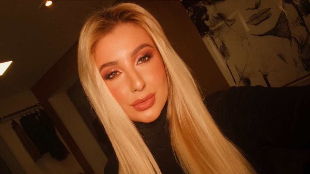 Jéssica de Paula posa para selfie em ambiente interno com iluminação quente usando roupa preta e cabelos loiros lisos soltos