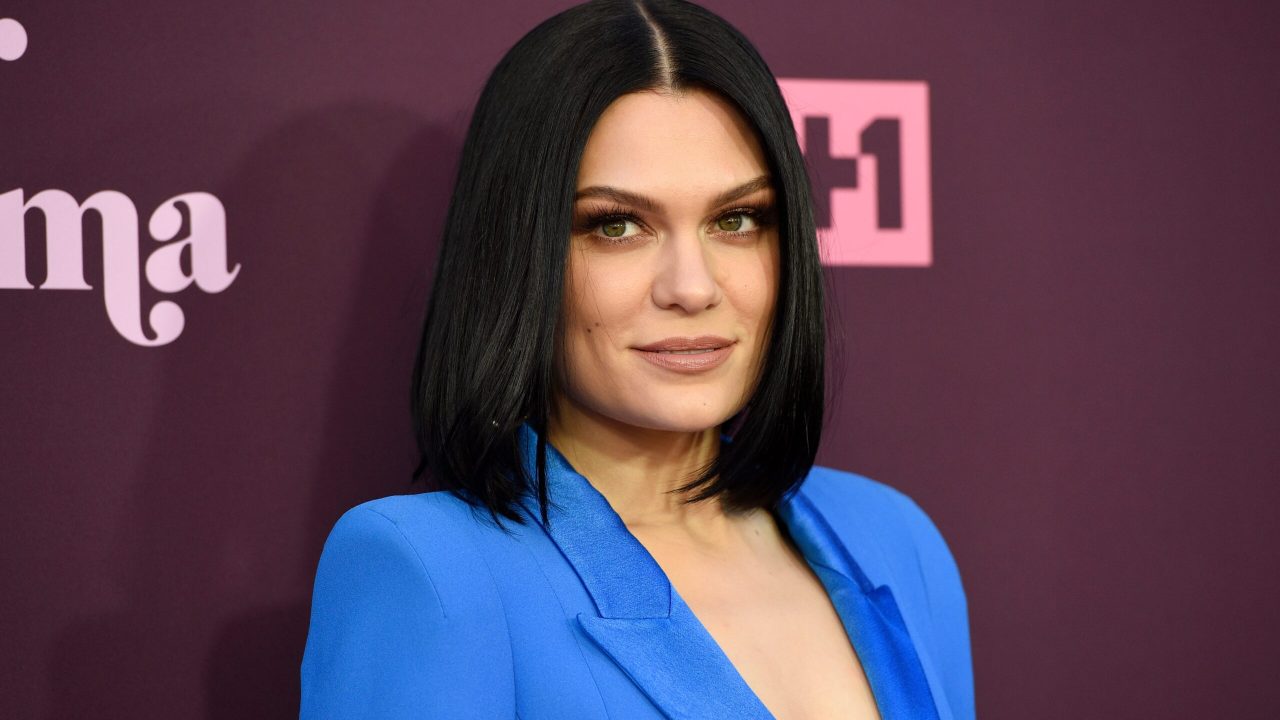 Jessie J posa sorridente em evento com fundo roxo e logotipo da VH1 ao fundo