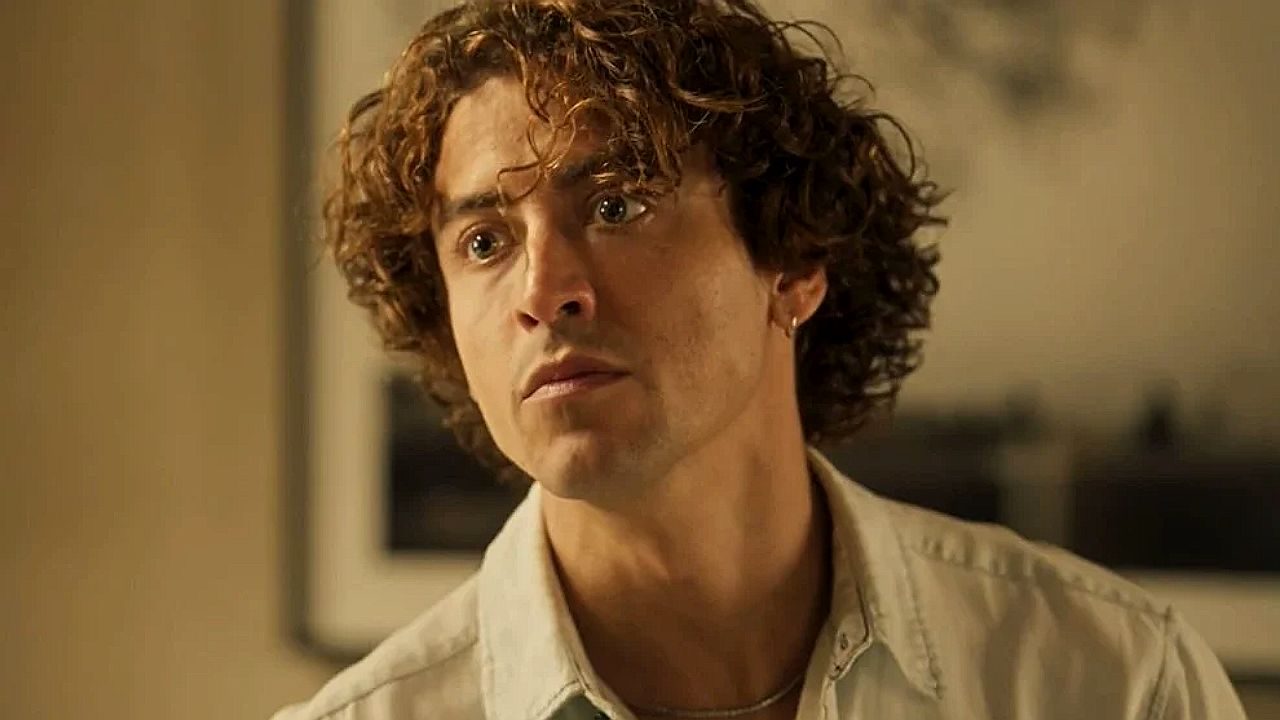 Homem jovem com cabelo cacheado olhando à frente com expressão séria, em um ambiente interno com iluminação quente.