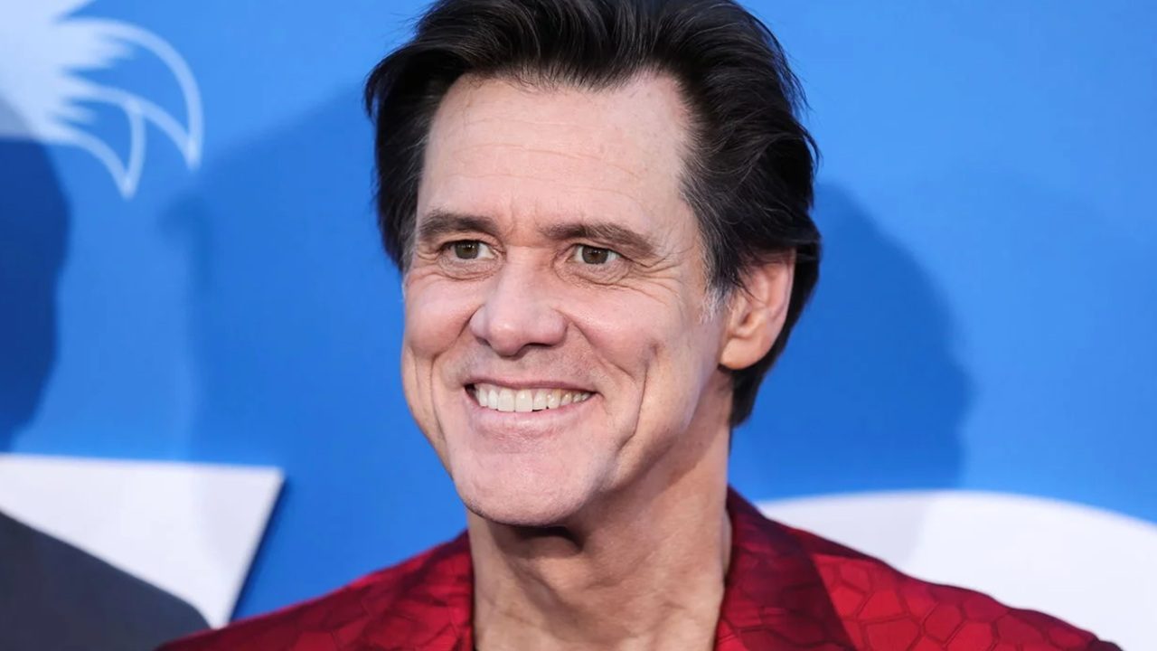 Homem sorrindo com cabelo escuro e jaqueta vermelha diante de fundo azul em evento público