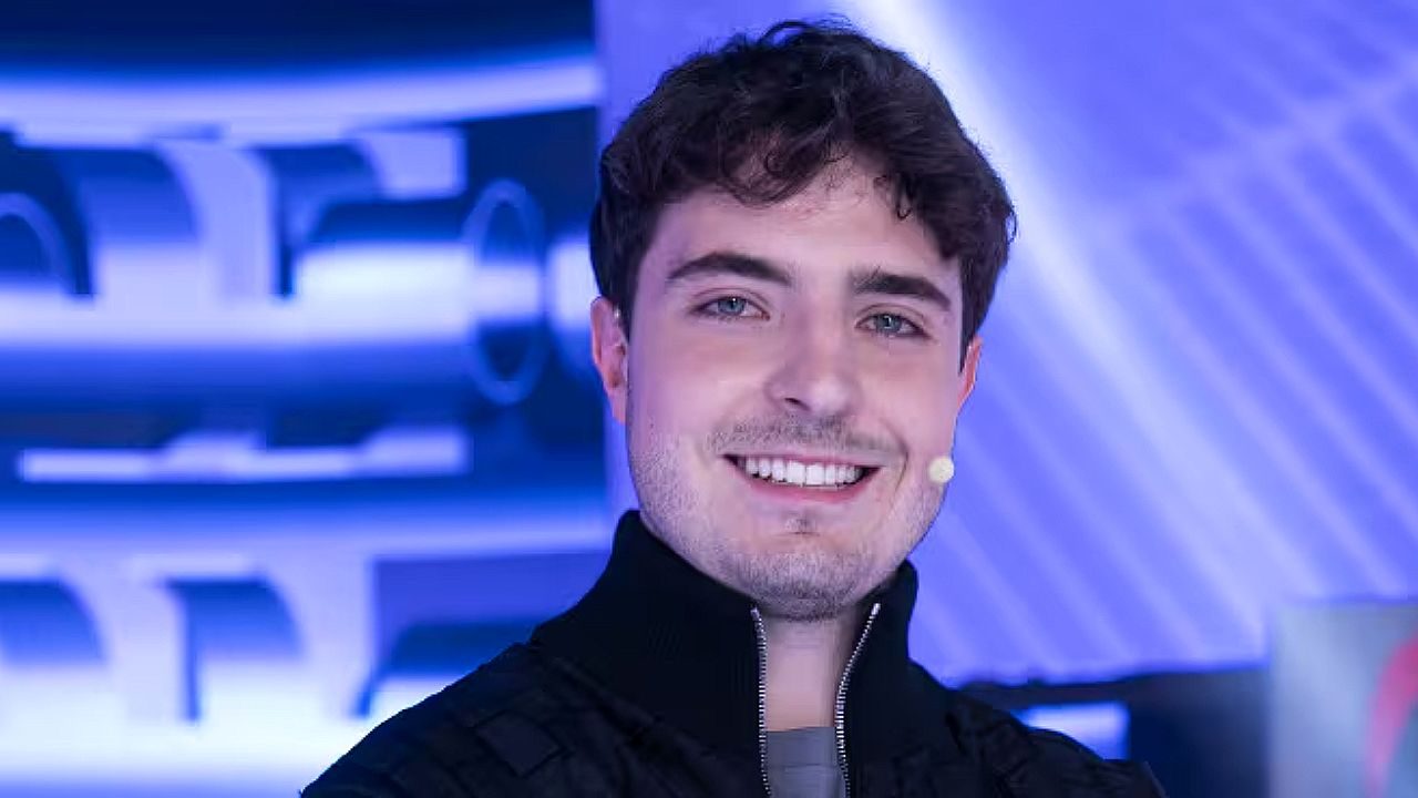 Homem jovem sorri durante participação em programa de televisão em estúdio com iluminação azul ao fundo e microfone de lapela no rosto