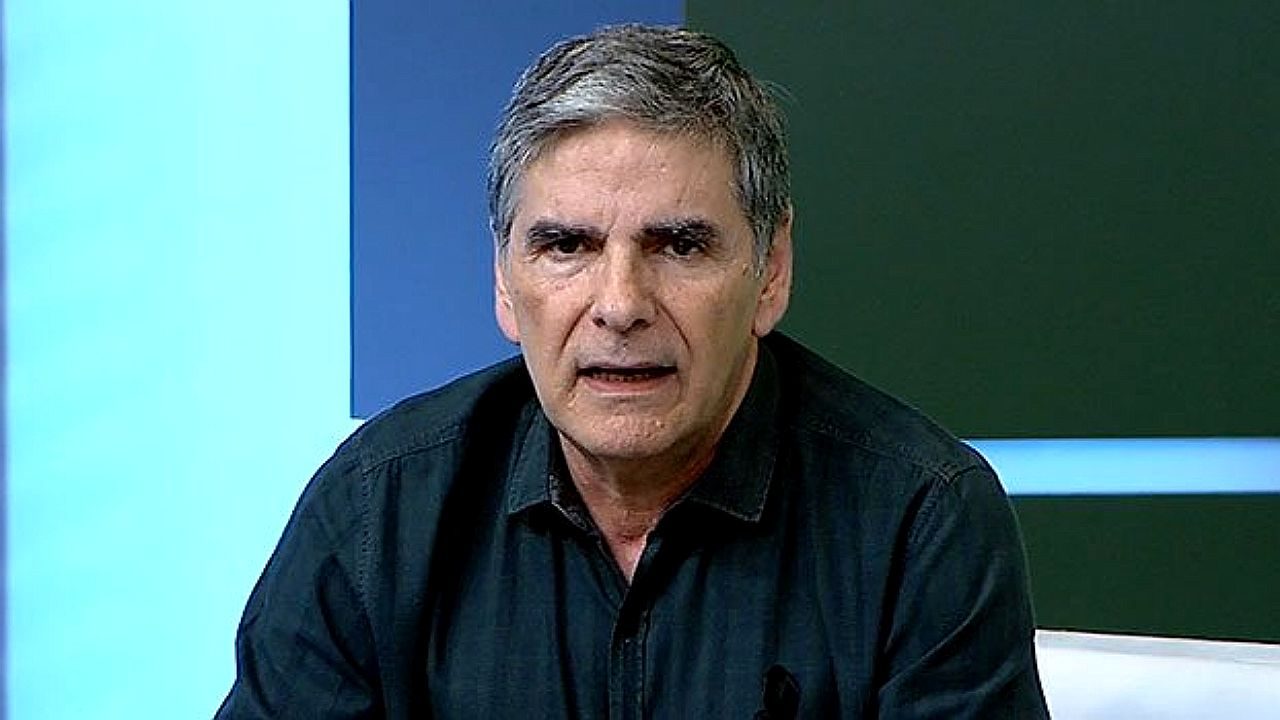 Homem de camisa preta fala sério durante apresentação em estúdio de telejornal com fundo em tons de azul e verde