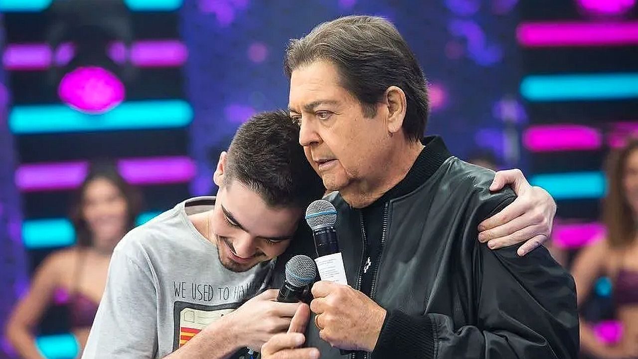 Apresentador abraça jovem emocionado enquanto segura microfone durante programa de auditório com plateia ao fundo