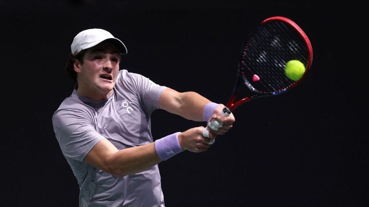 Jogador de tênis rebate uma bola com um backhand de duas mãos durante partida em quadra indoor, usando camiseta cinza, munhequeiras lilás e boné branco.