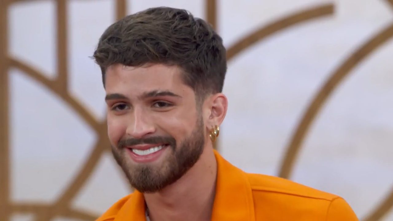 Homem jovem de barba bem aparada sorri enquanto olha para alguém fora de quadro; ele veste camisa laranja vibrante e usa pequenos brincos dourados, posando diante de um fundo claro com formas geométricas em tom dourado