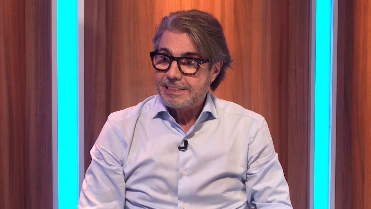 João Kleber sentado no estúdio durante participação no Programa Flávio Ricco com fundo de painéis de madeira iluminados