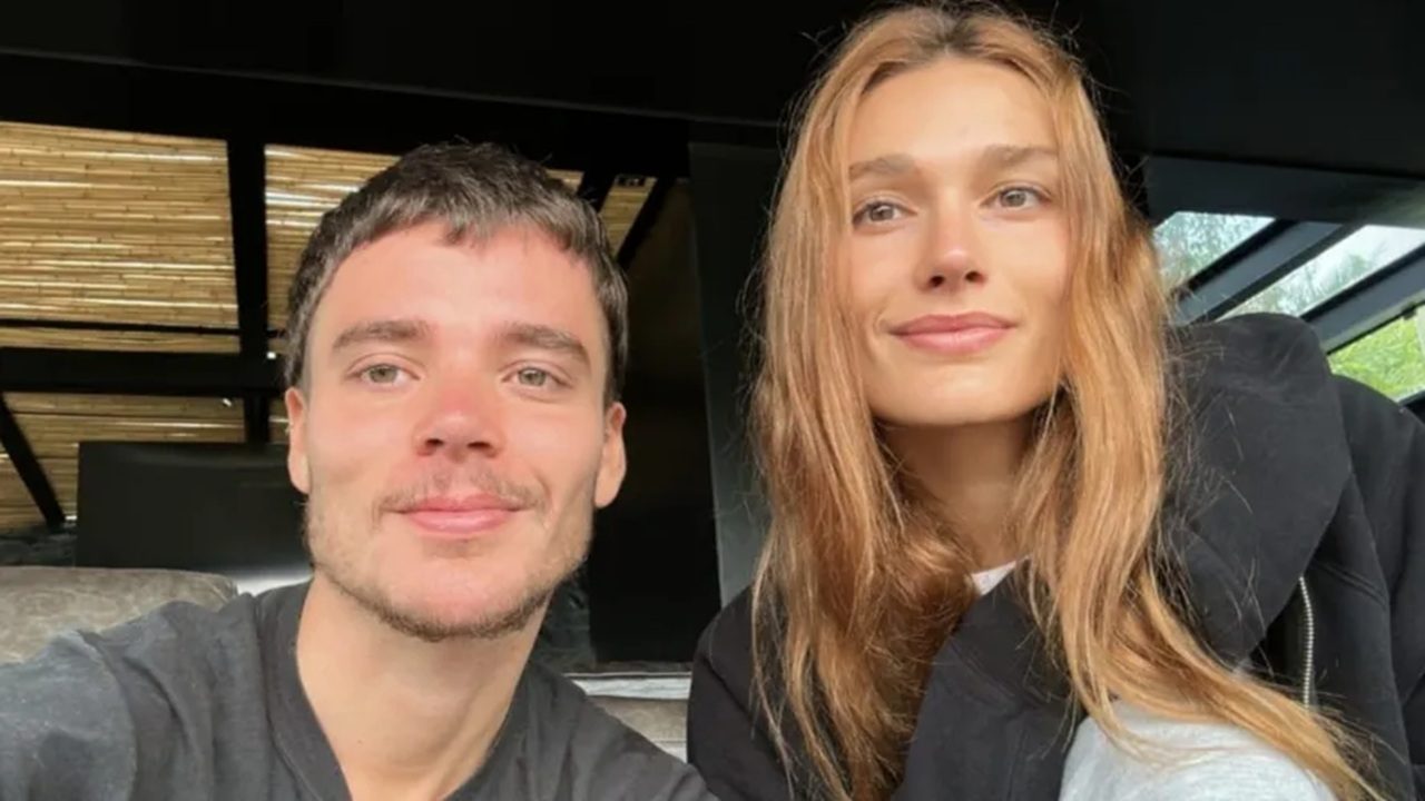 Duas pessoas jovens posam sorrindo para uma selfie em ambiente interno iluminado pela luz natural. À esquerda, um homem com cabelo curto castanho escuro e camiseta cinza; à direita, uma mulher de cabelos longos castanho-claros, usando casaco preto. Ao fundo, vê-se estrutura moderna com teto de ripas e janelas amplas