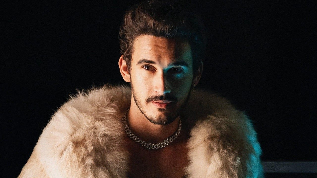 Filipe Bragança como João Raul posa com casaco de pele e colar em foto promocional da novela Coração Acelerado com fundo escuro