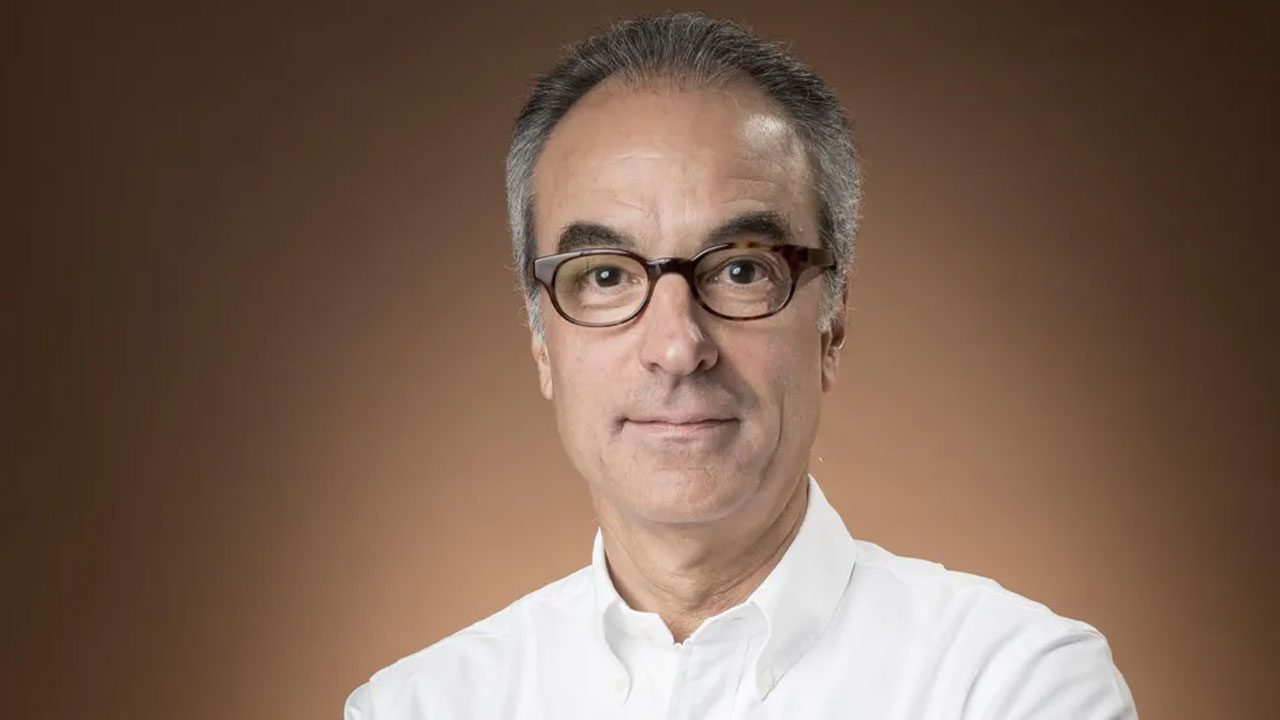 João Roberto Marinho usa camisa social branca e óculos de armação escura em foto com fundo marrom