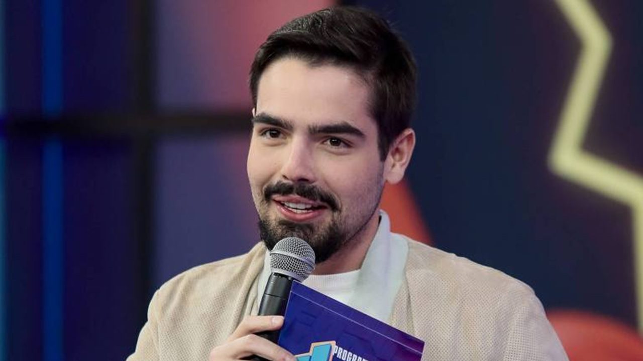 Apresentador segura microfone e cartão do programa enquanto fala em estúdio de atração de entretenimento na TV