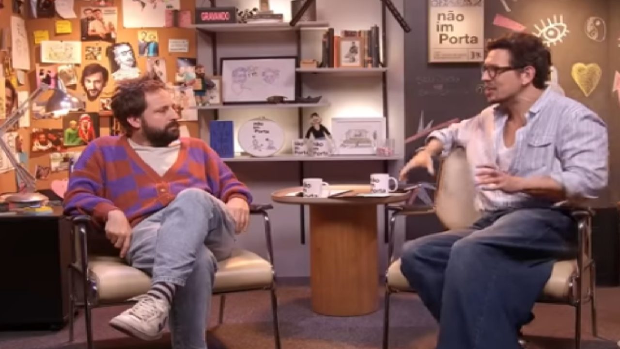 Gregório Duvivier e João Vicente de Castro durante conversa em cenário do programa, sentados em poltronas diante de uma mesa com canecas e decoração criativa ao fundo