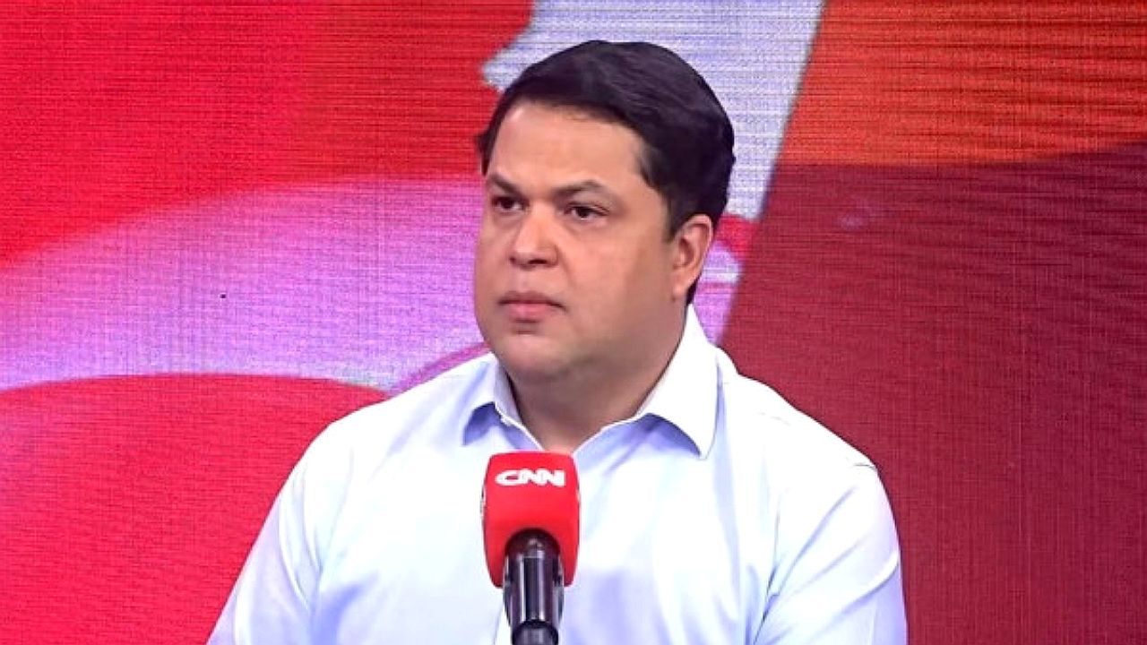 Um homem aparece em um estúdio de televisão, falando diante de um microfone com o logotipo da CNN Brasil. Ele veste camisa clara e tem expressão séria e concentrada, olhando levemente para o lado. Ao fundo, há um painel vermelho com elementos gráficos, característico de programas jornalísticos ou entrevistas.